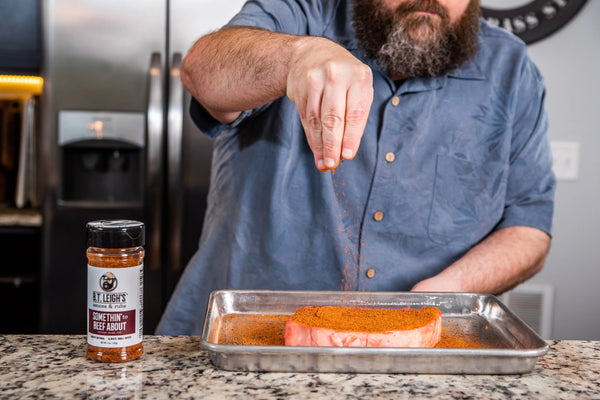 Man seasoning steak with B.T. Leigh's Somethin' to Beef About rub on a kitchen counter.