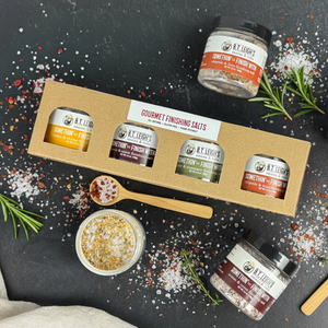 Collection of seasoning jars with a cardboard box on a dark surface with herbs and spoons.